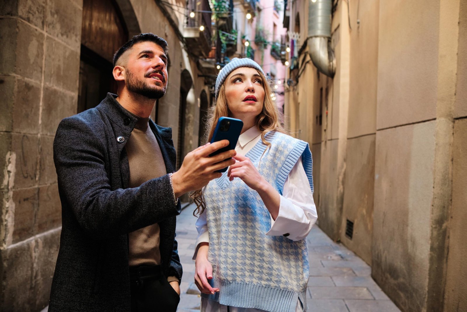 A man and a woman standing in a narrow, historic alleyway, looking up to navigate while checking a smartphone.