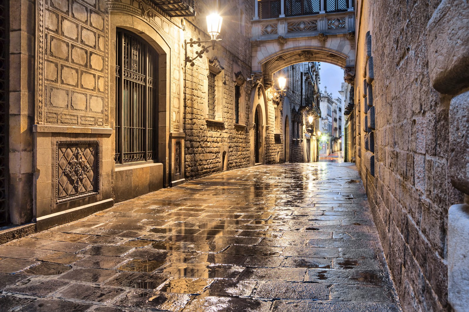 Narrow illuminated street in Barcelona’s Gothic Quarter with wet stone pavement and historic buildings at dusk